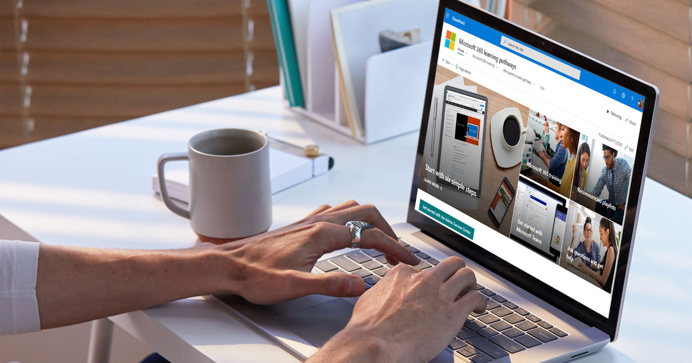 Image from https://adoption.microsoft.com/en-us/microsoft-365-learning-pathways/. Image of a white desk with a laptop open to Microsoft 365 Learning Pathways and a person's hands on the keyboard. Image also includes some other desk items like a cup of coffee and filed paperwork in the back. 